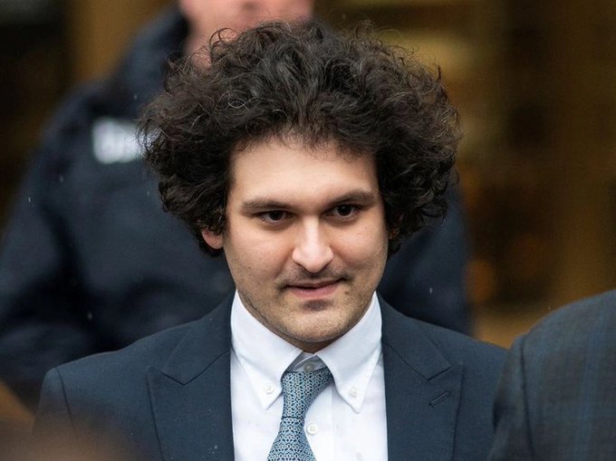 The image features a man with curly hair wearing a suit, tie, and jacket standing against a gray background. The man has his eyes closed while looking at the camera, giving him an intense gaze. There are two people partially visible behind him, one of whom is wearing a police uniform. The caption to the image reads "SBF is going to get pardoned isn't he chat. He should have to recover $LUNC as part of the agreement."