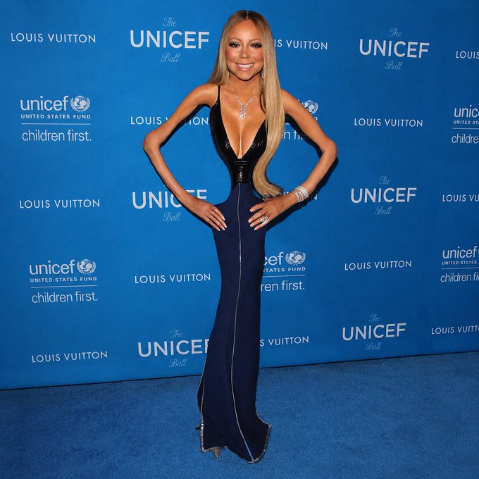 Mariah Carey poses in a black and navy blue gown on a blue carpet in front of a backdrop with logos for 'Louis Vuitton' and 'UNICEF Ball'.