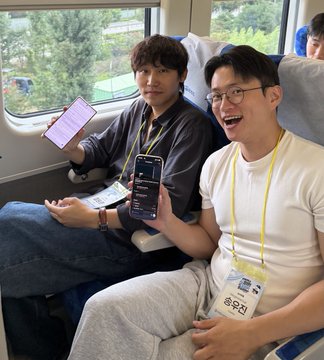 Two young men wearing lanyards sit on a train, smiling while holding up a phone displaying a code or development interface, capturing the casual, travel-friendly atmosphere of the SpaceX AI hackathon judging experience.