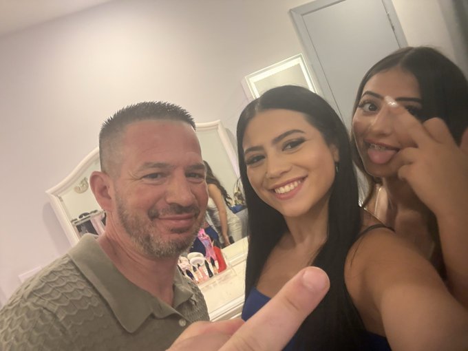 A selfie of three smiling people—a man on the left and two women on the right, with the women making peace signs—taken in what appears to be a bedroom or dressing room.