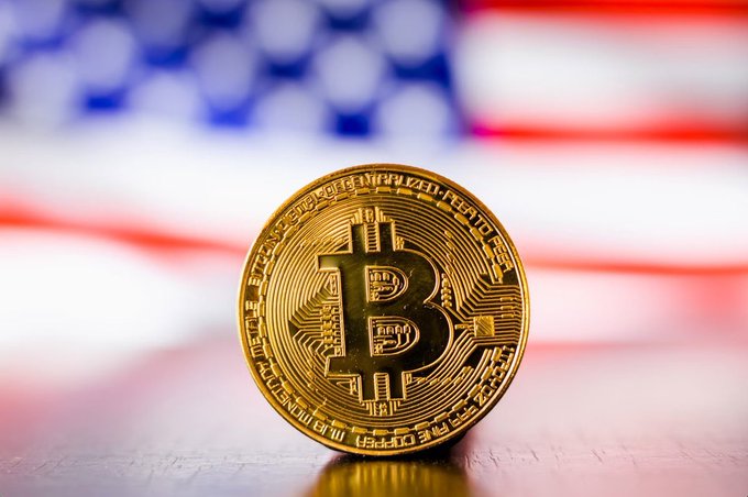 A physical Bitcoin coin stands upright against a blurred background of the American flag.