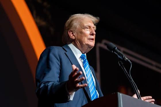 A man with blonde hair wearing a dark suit and blue striped tie speaks at a podium into a microphone, gesturing with his hand mid-address. The image accompanies the post about Trump's public statement on cryptocurrency, positioning him as the speaker making the remarks about America's leadership in Bitcoin and crypto ahead of a CFTC move.