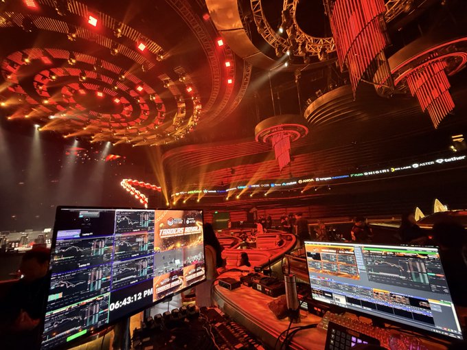 A vibrant nightclub or casino venue with elaborate red lighting, large ceiling chandeliers, and a central bar surrounded by multiple monitors displaying financial trading charts.