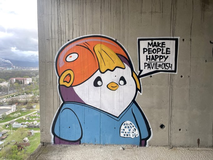 Street art/mural of a cartoon character with a cute face wearing a blue hoodie, with a speech bubble reading 'MAKE PEOPLE HAPPY PAVIL #0151', located in an urban setting with a view below.