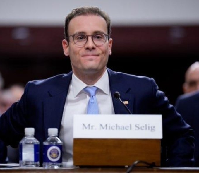 A man in a navy suit and blue tie sits at a desk with a nameplate reading 'Mr. Michael Selig', with two water bottles beside him at what appears to be a hearing.