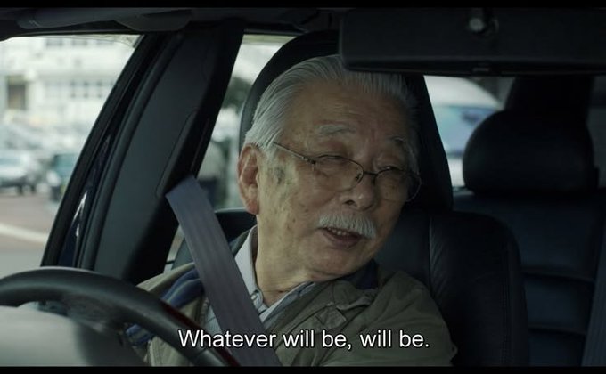 A scene showing an elderly Japanese man with white hair, mustache, and glasses sitting in a car with a seatbelt on, with the English subtitle 'Whatever will be, will be.'