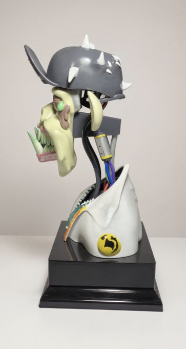 A bizarre sculptural figure features a spiked grey helmet, an elongated green face with sharp teeth, exposed mechanical neck cables, and a draped white cloth bearing a yellow circular logo, all mounted on a black base.