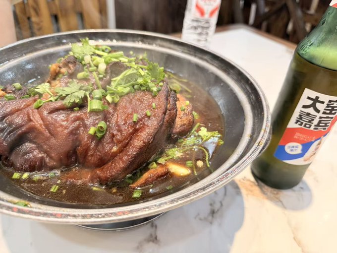 A large metal pot of braised meat (likely beef or pork trotters) garnished with cilantro and green onions, served with a bottle of DaBaiCai (大白蔡) beverage on the side.