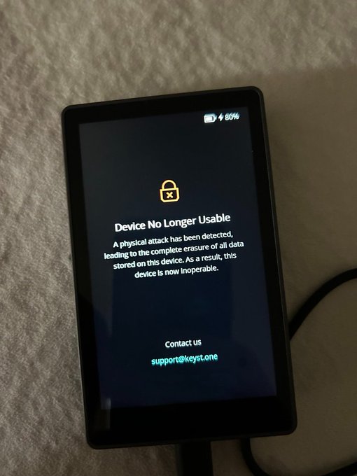 The image shows a black tablet or smartphone with a screen displaying an error message that reads "DEVICE NO LONGER USABLE" along with a lock symbol, indicating that the device has been disabled due to a security vulnerability. The device appears to be turned off and is placed on top of a bed.