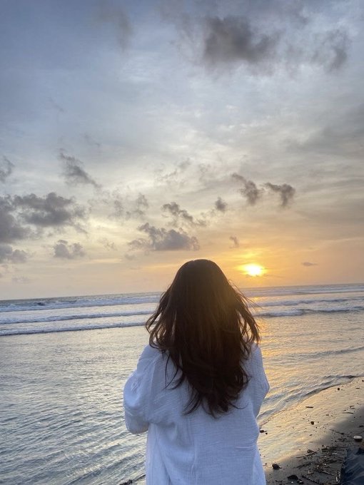 A woman with long dark hair, wearing a white linen shirt, stands on the shoreline gazing at a golden sunset over the ocean, its light reflecting across the rippling waves beneath a sky painted with soft clouds. The serene scene perfectly captures the post's reflection on nature's quiet wonders and invites viewers to pause and admire the simple beauty of a changing sky.
