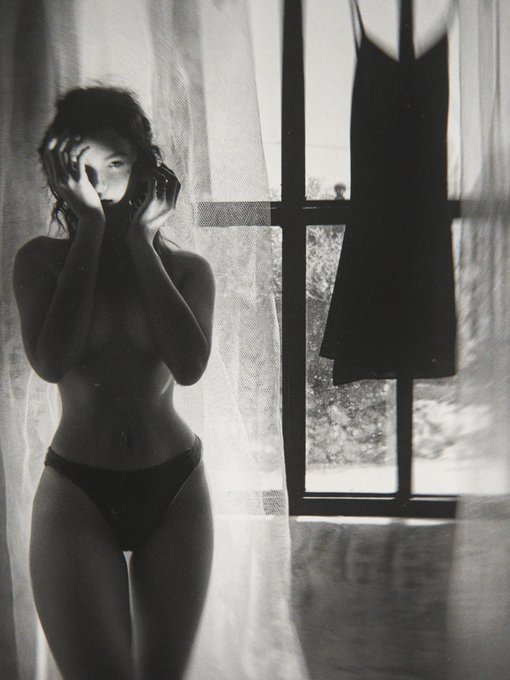 A black-and-white photograph titled "Portrait with a Dress" showing a woman in lingerie standing beside a sheer-curtained window, with a dark dress hanging prominently to her right, shared as an NFT on objkt.com (KT1FjGN) for #TezosTuesday.