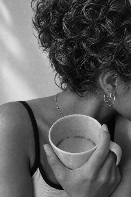 A black and white photograph showing a close-up of a person with short curly hair and hoop earrings, holding a ceramic cup near their shoulder while wearing a spaghetti-strap top.