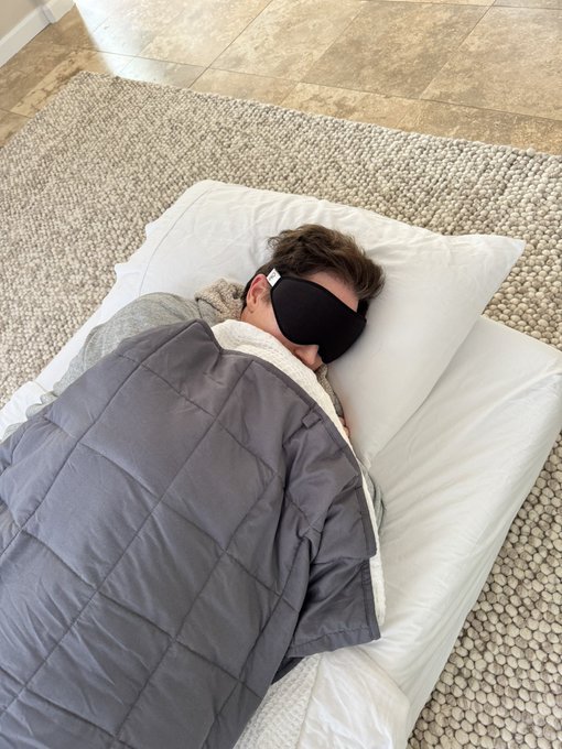 The image shows a person laying down with their eyes closed while wearing a black eye mask. The person appears to be resting or sleeping peacefully. In the background, there are two pillows placed against the wall, one of which has a pillowcase that matches the color of the eye mask. The text in the image reads: "I sleep with peace of mind knowing I can send money anywhere in the world instantly using Polygon PoS payments. Fast, secure and borderless, that's the power of Web3 transactions." This message is related to the person wearing an eye mask as it suggests a connection between the comfort provided by the eye mask and the convenience offered by modern financial services through online platforms like Polygon payments.
