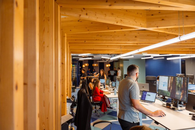 A team collaborates in a modern, wood-accented office space with exposed beam ceilings and multiple workstations, where employees work on dual monitors under warm overhead lighting. The image reflects the post's message of teamwork and shared purpose at xMoney, showing colleagues actively engaged together in a unified work environment.