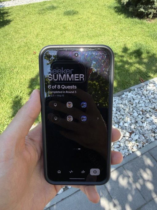 A person's hand holding a smartphone outdoors over grass and gravel, displaying a "seeker SUMMER" event screen showing "6 of 8 Quests Completed in Round 3" with several profile icons.