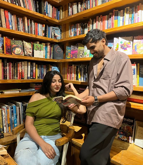 A man teaches a woman how to read a book in a bookstore, with titles like "Ruskin Bond" and "The Ring" visible on the shelves, playfully illustrating the post's joke about launching a "paid course" to teach trading and avoiding liquidations.