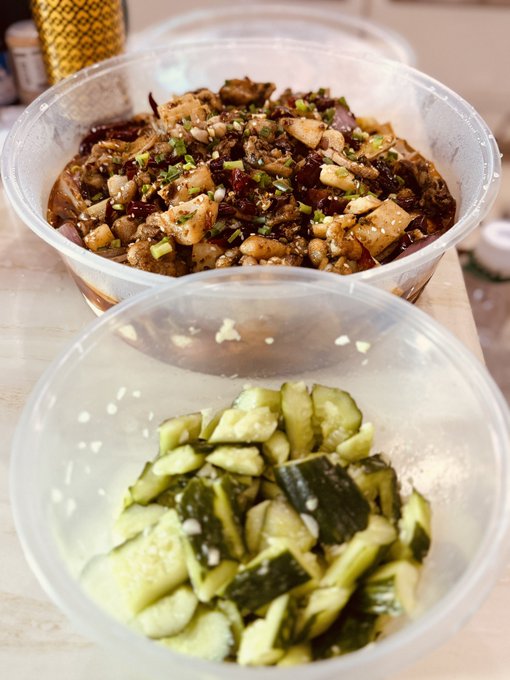 Two plastic containers holding a spicy stir-fry dish topped with sesame seeds and chives, alongside a smashed cucumber salad.