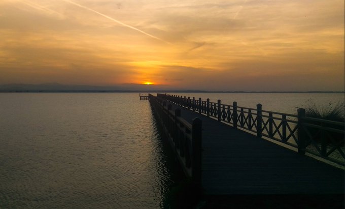 A photograph of a sunset over a calm sea, with a silhouetted wooden pier extending into the water and faint mountain ranges visible on the horizon.