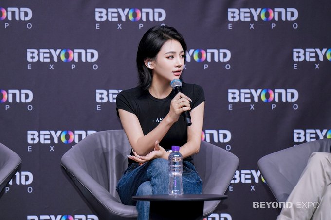 The image features a woman sitting in a chair, holding a microphone as she speaks into it. She appears to be at an event or gathering where she is addressing an audience. The background of the image shows a black wall with white text that reads "Beyond Expo". There are also chairs and a water bottle visible in the scene.