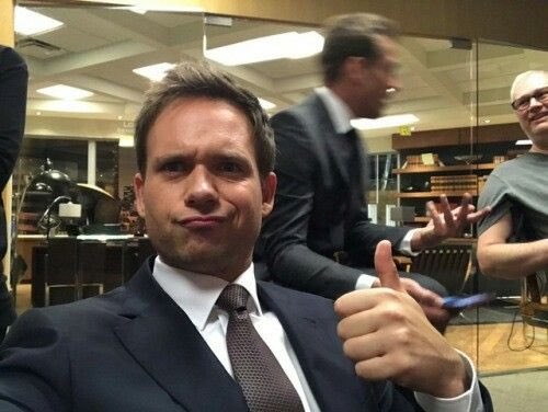 A selfie of actor Joel McHale in a dark suit and tie giving a thumbs-up in what appears to be an office setting.