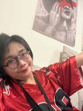 A selfie of a smiling person with glasses wearing a red patterned shirt, with a framed black-and-white photo of a screaming person (with a red censor bar over their eyes) hanging on the wall behind them.