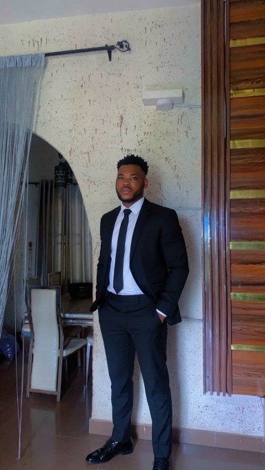 A young man in a black suit, white shirt, and black tie posing indoors against a textured wall next to a wooden door and an arched mirror.