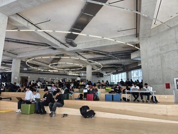 The image shows a group of people sitting at tables with laptops open, working or studying together in an indoor space. There are several individuals scattered throughout the room, some sitting alone while others collaborate. The scene takes place within a large building, possibly a library or a common area designed for group work and study sessions. The atmosphere appears to be focused and productive as people engage in their tasks at hand.