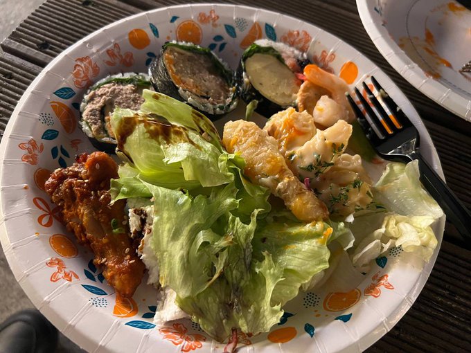 A paper plate holds an outdoor meal featuring green leaf lettuce, fried chicken, shrimp, and seaweed-wrapped rolls, with a black plastic fork resting on the side.