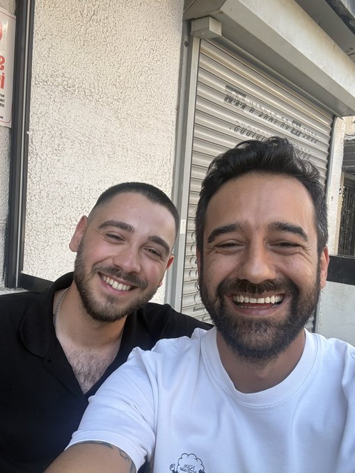 Two smiling men pose for a selfie in front of a closed metal storefront, with the bearded man on the right in a white t-shirt appearing to take the photo alongside his friend from Chainlink Turkey.