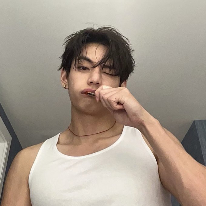 A young man wearing a white tank top takes a selfie while brushing his teeth with one eye closed.