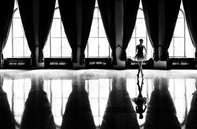 A black and white photograph of a ballerina in a tutu standing in a grand hall with large windows, her reflection visible on the polished marble floor.