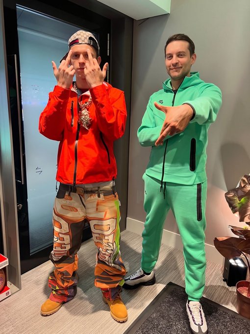 Image 9: Two men posing indoors; the man on the left wears a bright orange jacket, patterned pants, and holds up his middle fingers, while the man on the right wears a bright green tracksuit and extends his hand outward.