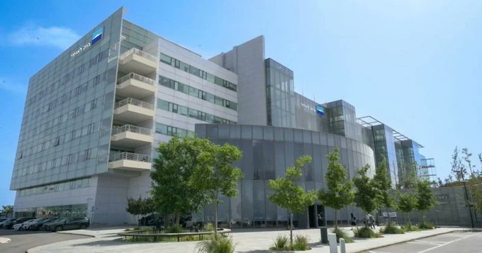 A modern, multi-story office building with glass facades, featuring the 'WILLY.FM' logo, situated on a paved plaza with small trees.
