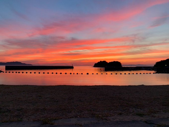 A scenic sunset view over a calm body of water with vibrant pink and orange clouds reflecting on the surface, framed by silhouetted mountains and a stone breakwater with floating buoys.