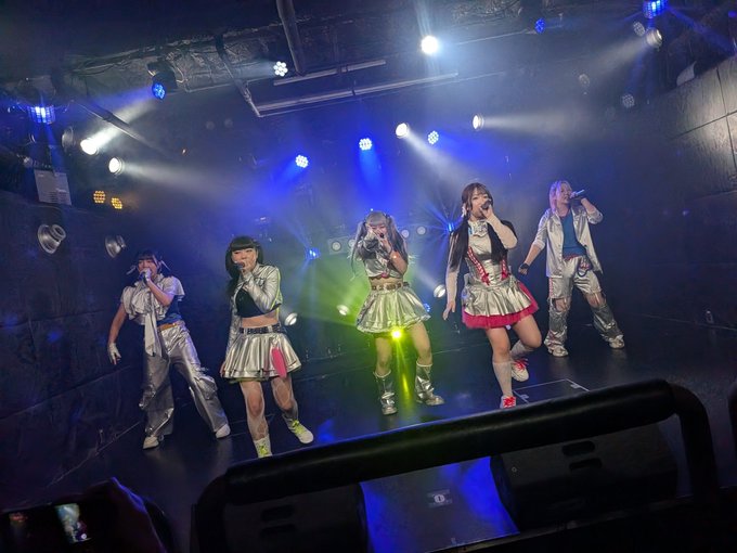A J-pop or J-idol group performs on a dimly lit stage with blue and green lighting, featuring five members in shiny metallic silver outfits with colorful accents, singing into microphones.