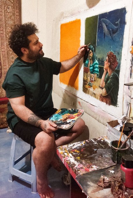A man sits on a small stool in an art studio, painting a colorful canvas with a palette in hand; the painting shows figures at a table, a window, an orange wall section, and a whale visible outside.