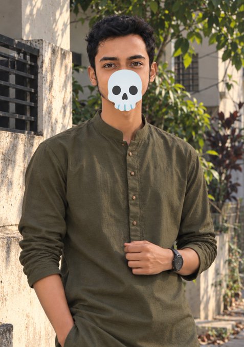 A young man in an olive green kurta stands in front of foliage, wearing a digital skull sticker over his face.