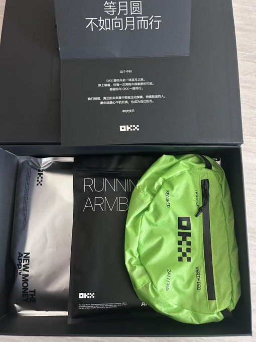 An unboxing photo showing a black gift box containing items including a silver 'NEW MONEY' package, a black booklet with Chinese text reading '等月圆 不如向月而行', and a bright green 'RUNNING ARMB' (ARMOUR) fanny pack with '24.11.2555' printed on it.