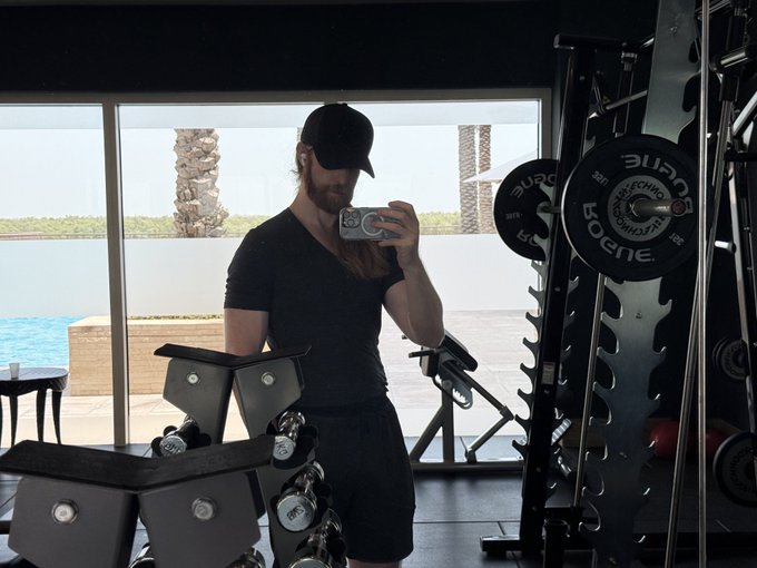 A mirror selfie of a bearded man wearing a black cap and t-shirt working out in a gym, with Rogue weight plates visible on the right.