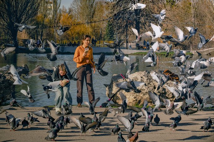 A photograph of a woman and child at a park surrounded by a large flock of pigeons in flight, with the woman holding what appears to be bird feed in an orange sweater.