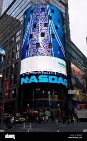 The image shows a large blue and white building with a prominent sign that reads "Nasdaq" at its top, indicating that this is an important financial center or stock exchange. The street below has several people walking around, some of whom are carrying backpacks. There are also multiple cars parked along the street, as well as traffic lights controlling the flow of vehicles and pedestrians. A person can be seen standing near a lamppost on the sidewalk.