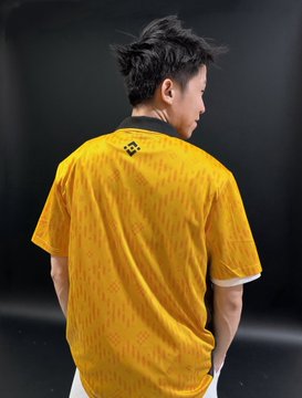 The image features a man wearing a yellow shirt with black stripes, standing against a black background. The shirt has a unique design that includes a pattern of red dots on a white background. The man appears to be posing for the photo while looking at the camera. The image also contains text that reads "感謝 @binancezh 寄來的限量Team Binance足球球衣".