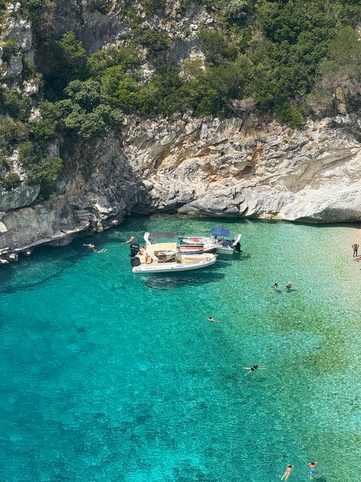 An aerial view of a small boat anchored in crystal-clear turquoise waters near a rocky cliff, with several people swimming around it.
