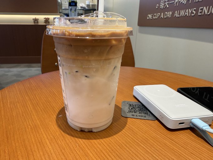 An iced coffee drink in a clear plastic cup sits on a wooden table next to a white power bank charging a phone, with a partial sign reading 'ONE CUP A DAY ALWAYS ENJOY' in the background.