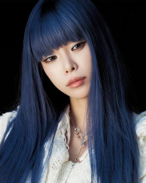A young woman with long, dark blue hair and bangs gazes at the camera against a black background, wearing a cream-colored knit top and silver necklaces. The ethereal, striking image seems to evoke the sleek, crypto aesthetic associated with the "Hays Ethereum Re-entry $ETH" trading post, using her bold styling to visually capture attention for the Ethereum announcement.