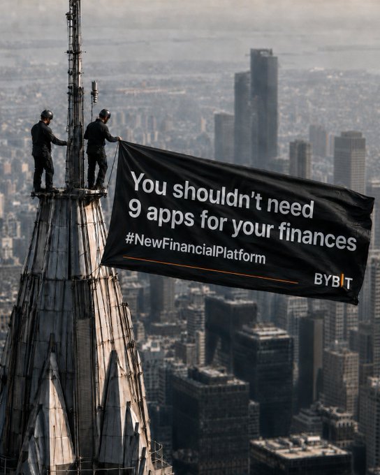 Two workers stand atop a tall building spire holding a large black banner for Bybit that reads "You shouldn't need 9 apps for your finances #NewFinancialPlatform."