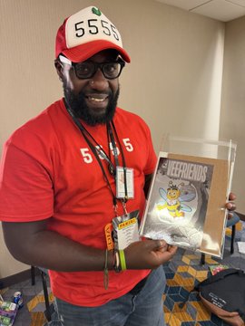 A man wearing a red "5555" cap and matching red shirt smiling while holding a packaged comic book or collectible titled "DEEFIENDS" at what appears to be a convention.