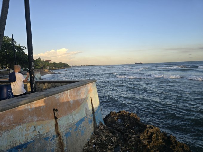A scenic view of the ocean from a weathered, painted concrete wall, with a person standing nearby and cargo ships visible in the distance under a clear sky.