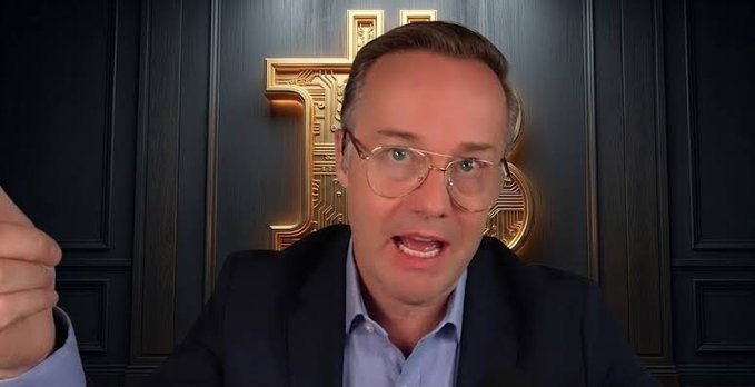 A man with glasses wearing a dark suit jacket and light blue shirt speaks directly to the camera, with a gold Bitcoin-style logo visible on the dark wall behind him.
