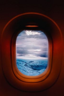 A view through an airplane window showing snow-covered mountains and a cloudy sky, with the window frame glowing orange in the dark cabin.